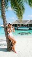 Young woman, Caucasian ethnicity, wearing white swimwear and sun hat, photographing tropical beach resort with overwater bungalows and turquoise lagoon waters.