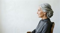 Elegant senior woman with gray hair in braided updo, wearing dark clothing, seated in profile against white background, suitable for healthcare, wellness, aging, and lifestyle editorial use.