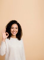 Young woman with curly dark hair and Latin ethnicity wearing cream sweater making okay hand gesture against beige background, expressing approval and satisfaction for lifestyle and business concepts.