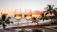 Two glasses of white wine on tropical beach table at sunset with palm trees and ocean view for romantic dining and vacation relaxation.