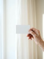 Senior male hand holding blank white card against neutral beige curtain background. Mockup for business cards, invitations, or promotional materials in bright interior setting.