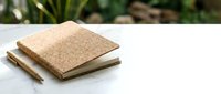 Cork notebook with wooden pen on white marble surface with blurred green foliage background, suitable for stationery and office design projects.