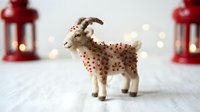 Handmade felt goat figurine with red polka dots, standing on white wooden surface with festive red lanterns and warm bokeh lights in background for holiday decoration.