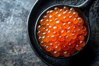 Fresh red caviar in black ceramic spoon on dark textured background, bright orange salmon roe spheres with natural gloss, macro food photography.