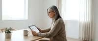 Mature woman with gray hair wearing glasses and beige sweater using tablet computer at wooden desk in bright modern home office workspace.