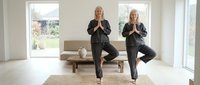 Senior women practicing yoga in bright modern living room, performing tree pose together for fitness and wellness routine.