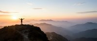 Person with arms raised celebrating success on mountain peak at sunrise with layered hills and golden sky creating inspirational achievement moment.