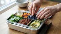 Caucasian hands reaching into a compartmentalized meal prep container filled with fresh vegetables, grains, berries and protein portions on wooden surface near window for healthy eating planning.