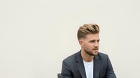Young adult male with light brown hair and facial hair wearing a dark gray blazer over a white shirt, isolated on white background for design and commercial use.