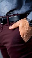 Close-up of male hand in pocket of burgundy trousers with black leather belt, wearing formal business attire. Cropped view showing refined menswear details.