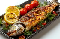 Grilled whole fish with charred lemon, fresh cherry tomatoes and dill garnish served on dark plate. Close up view of Mediterranean style seafood dish.