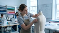 Young Caucasian female fashion designer working on dress form mannequin in bright modern studio workspace with colleagues in background.