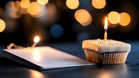 Single cupcake with lit birthday candle next to open blank book against blurred warm bokeh lights creating magical celebratory atmosphere.