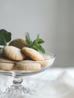 Fresh shortbread cookies served on elegant glass cake stand with mint leaves garnish against light background, minimalist food photography styling.