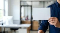 Male professional in dark blue shirt holding blank white card in modern office environment with blurred background, suitable for business messaging and corporate communications.