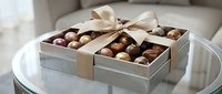 Luxury chocolate assortment in elegant silver box with gold ribbon bow, displayed on white round table for gifting and celebrations.