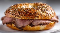 Freshly baked bagel with everything seasoning filled with tender sliced roast beef, served on white plate. Perfect for breakfast or lunch menu.
