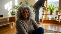 Happy African American woman with curly gray hair sitting on floor in bright living room with plants while partner tends to greenery in background.