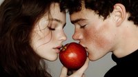 Young romantic couple sharing red apple, intimate close up portrait against gray background showing temptation and desire in contemporary style.