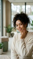 Young woman with curly dark hair and medium brown skin tone wearing cream cardigan, smiling while touching face in bright modern home interior with plants and windows, lifestyle portrait.