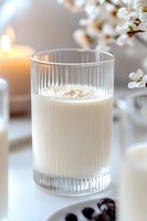 Fresh milk in ribbed glass with spring cherry blossoms and candle in soft morning light, creating serene breakfast atmosphere on white table surface.