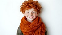 Happy redhead child with curly hair wearing orange knitted scarf smiling at camera on white background. Cozy winter fashion portrait for kids clothing.