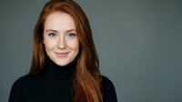 Young Caucasian woman with long red hair wearing black turtleneck sweater smiling at camera against gray background for professional headshot portrait.