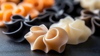 Colorful pasta shells in orange, black, and white arranged on dark background, showing delicate curves and textures of fresh homemade Italian farfalle noodles.