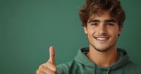 Young man in green hoodie showing thumbs up gesture against green background with confident smile and positive attitude, expressing approval and success.