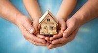 Family hands protecting wooden house model symbolizing home insurance, real estate investment and property protection concept on blue background.