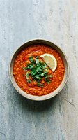 Hearty red lentil soup with fresh herbs and lemon slice in rustic ceramic bowl on textured stone surface for healthy comfort food recipes.