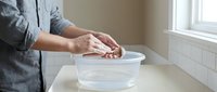 Woman in gray apron kneading dough in white ceramic bowl on kitchen counter near window. Hands working with bread dough for homemade baking and cooking.