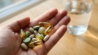 Hand holding golden supplement capsules and pills with glass of water on wooden surface for wellness routine.