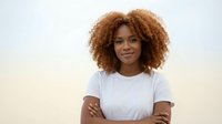 Young African American woman with curly hair smiling confidently with crossed arms wearing white t-shirt against soft blurred background for lifestyle content.