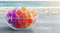 Colorful fruit gummy candies in glass bowl on wooden table with blurred ocean beach background. Sweet treats for summer vacation dessert concept.
