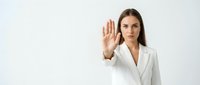 Young professional businesswoman in white blazer making stop gesture with raised palm against neutral background for corporate communication concepts.
