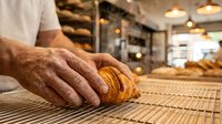Baker's hand holding fresh croissant on cooling rack in bakery shop. Artisan bread production and pastry making for cafes, restaurants, and food service businesses.