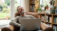 Mature Caucasian man with beard and glasses relaxing in leather armchair with laptop in bright home library. Remote work, freelancing, retirement lifestyle.