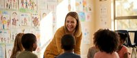 Young female teacher with blonde hair smiling while interacting with diverse elementary school children in bright classroom with educational posters on walls.