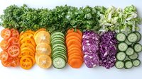 Fresh sliced vegetables and citrus arranged in colorful rows including carrots, oranges, cucumber, red cabbage, lettuce and parsley on white background.