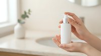 Close-up of hands holding white cosmetic spray bottle in bright bathroom setting with natural light and potted plant, suitable for skincare and beauty product marketing.