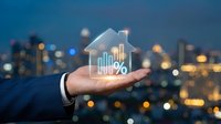 Businessman hand holding glowing house icon with percentage symbol and charts over blurred city lights background representing real estate investment.