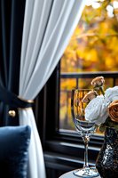 Empty wine glass near window with white roses and autumn foliage view, elegant navy curtains and velvet cushion create cozy romantic atmosphere.