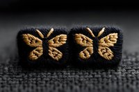 Delicate golden butterfly embroidery on black fabric buttons creates elegant detail against textured background. Macro photography captures intricate needlework pattern.