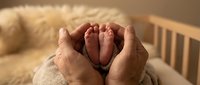 Adult hands forming heart shape around newborn baby feet in crib, expressing love and parental care for new family member.