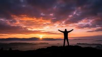 Person silhouette with raised arms celebrating success at mountain summit during dramatic sunrise with colorful sky and valley below for motivation concepts.