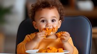 Mixed race toddler in orange shirt eating messy chicken wings with sauce-covered face, sitting in dining chair showing authentic childhood mealtime moment.