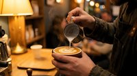 Skilled barista pouring steamed milk into a latte, creating intricate latte art in a cozy coffee shop with warm ambient lighting.