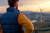 Man in blue and yellow jacket observing wind turbines at sunset from elevated viewpoint, renewable energy landscape concept for sustainability and clean power generation.