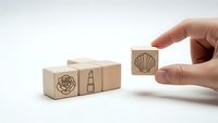 Hand placing wooden cube with seashell icon next to blocks featuring rose and lipstick symbols on white background for branding and design concepts.